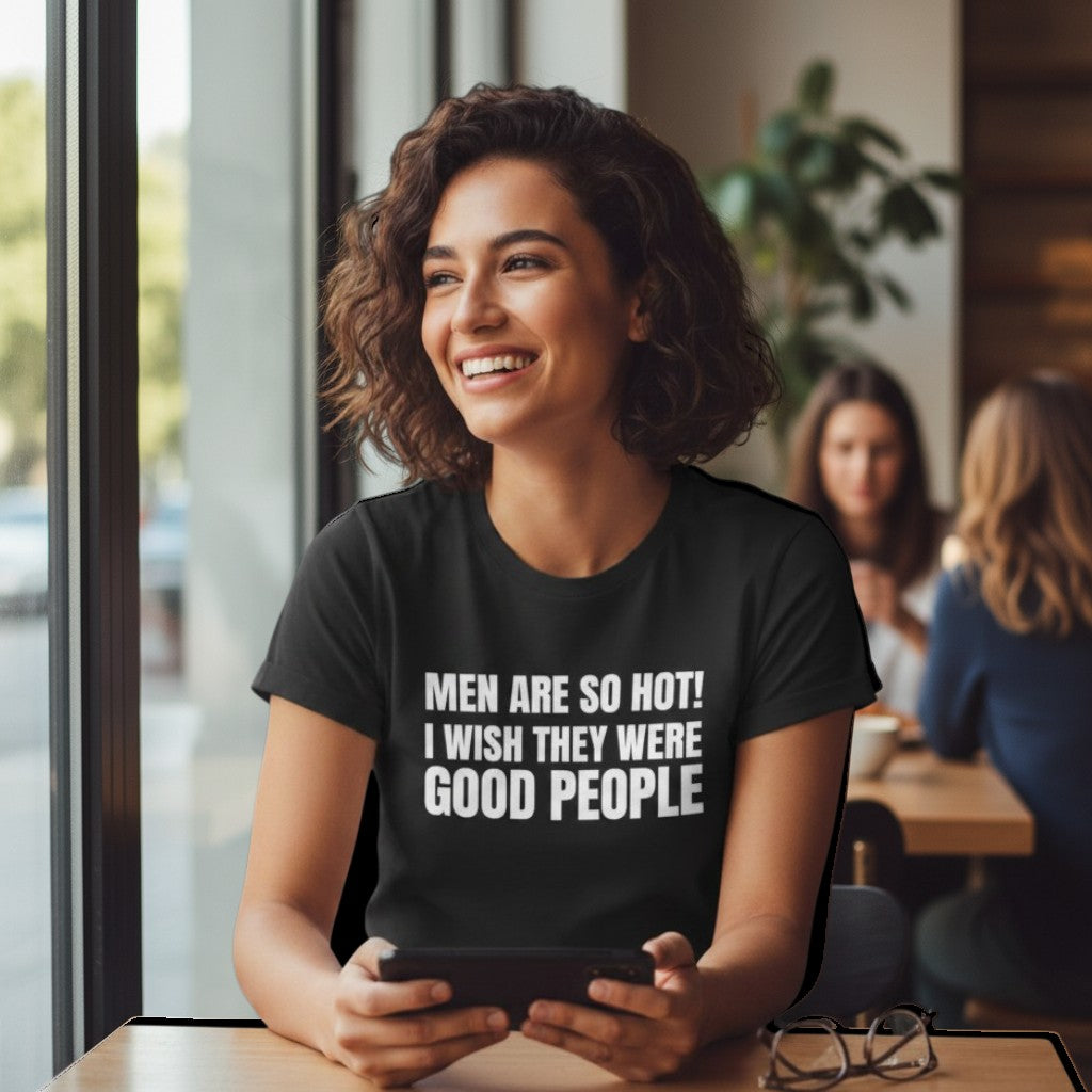 Eine Frau mit lockigem Haar trägt ein schwarzes spiritualsavagee T-Shirt mit dem Aufdruck MEN ARE SO HOT I WISH THEY WERE GOOD PEOPLE in einem Café.